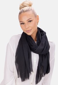 Écharpe légère noire en tissu semi-transparent avec une texture froissée. Les bords sont légèrement effilochés, portée drapée autour d'une chemise blanche.