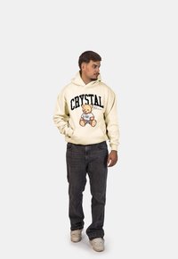 Felpa crema con testo "CRYSTAL Paris" e grafica di un orso; abbinata a jeans scuri e sneakers bianche. Stile streetwear casual.