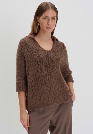 Brauner gestrickter Pullover mit V-Ausschnitt und umgeschlagenen Manschetten. Mit einem strukturierten, groben Design. Kombiniert mit neutral gefärbten Hosen.