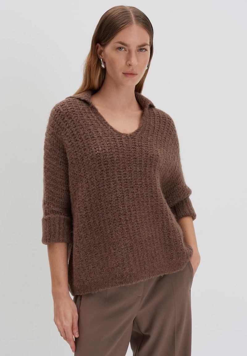 Brauner gestrickter Pullover mit V-Ausschnitt und umgeschlagenen Manschetten. Mit einem strukturierten, groben Design. Kombiniert mit neutral gefärbten Hosen.