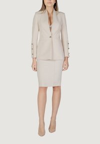 Blazer beige su misura con un unico bottone, bavero deciso e maniche lunghe con bottoni dorati. Abbinato a una gonna a matita beige aderente.
