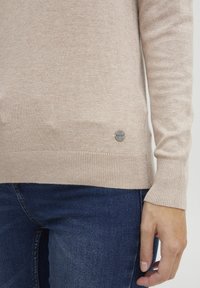 Bežani pleteni džemper s rebrastim ručnim i donjim rubovima. Ima mali okrugli logotip pri dnu. Nosite s plavim trapericama.