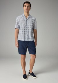 Kortærmet, knap-op skjorte med et blåt og hvidt geometrisk mønster, sammen med marineblå shorts og marineblå slip-on sko.