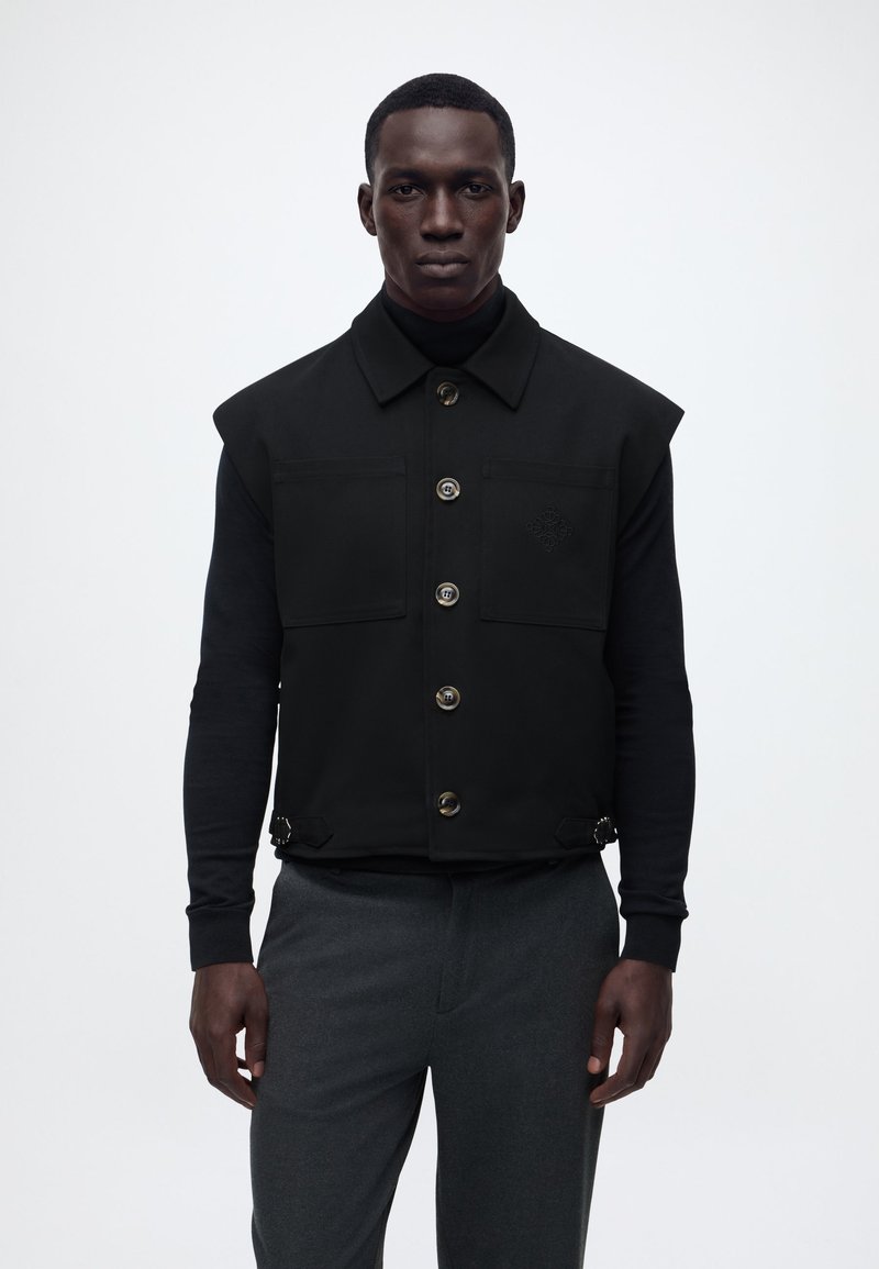 Άνδρας που φοράει ένα μαύρο γιλέκο με κουμπιά πάνω από μαύρο μακρυμάνικο μπλουζάκι και γκρι παντελόνι, στέκεται μπροστά σε λευκό απλό φόντο.