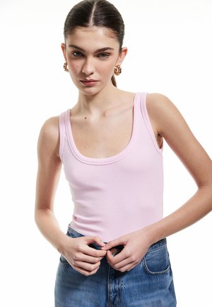 Lichtroze geribbelde tanktop met brede bandjes, gecombineerd met hoge taille blauwe spijkerbroek. Model raakt de knoop van de spijkerbroek aan.