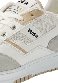 Baltas sneakers, kas izgatavoti no teksturēta kanvas un zamšādās, ar gaiši brūniem akcentiem un melnu "MoEa" zīmolu sānos.