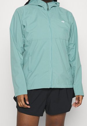 Personne portant une veste à capuche vert clair avec une fermeture éclair à l'avant et un short noir, debout devant un fond uni.