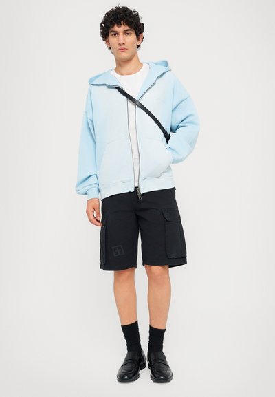 Helle blaue Zip-Hoodie, schwarze Cargo-Shorts mit Seitentaschen und schwarze Slipper. Darunter ein schlichtes weißes T-Shirt, getragen mit schwarzen Socken.