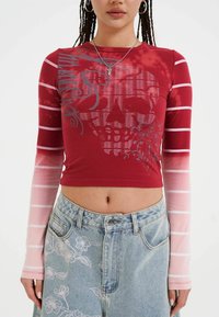 Červený crop top s dlouhým rukávem a grafikou lebky, se růžovými pruhovanými rukávy. V kombinaci s modrou džínovou sukní s vysokým pasem, zdobenou vyšívanými květinami.