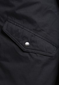 Poche de veste en tissu noir avec rabat incliné fermé par un bouton-pression rond en argent gravé du texte "PIER ONE".