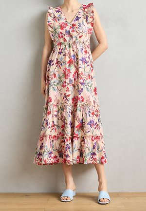 Person iført en ærmeløs, blomsterprintet midi kjole med flæser og lyseblå åbentåede blokhælsandaler, der står mod en neutral væg.