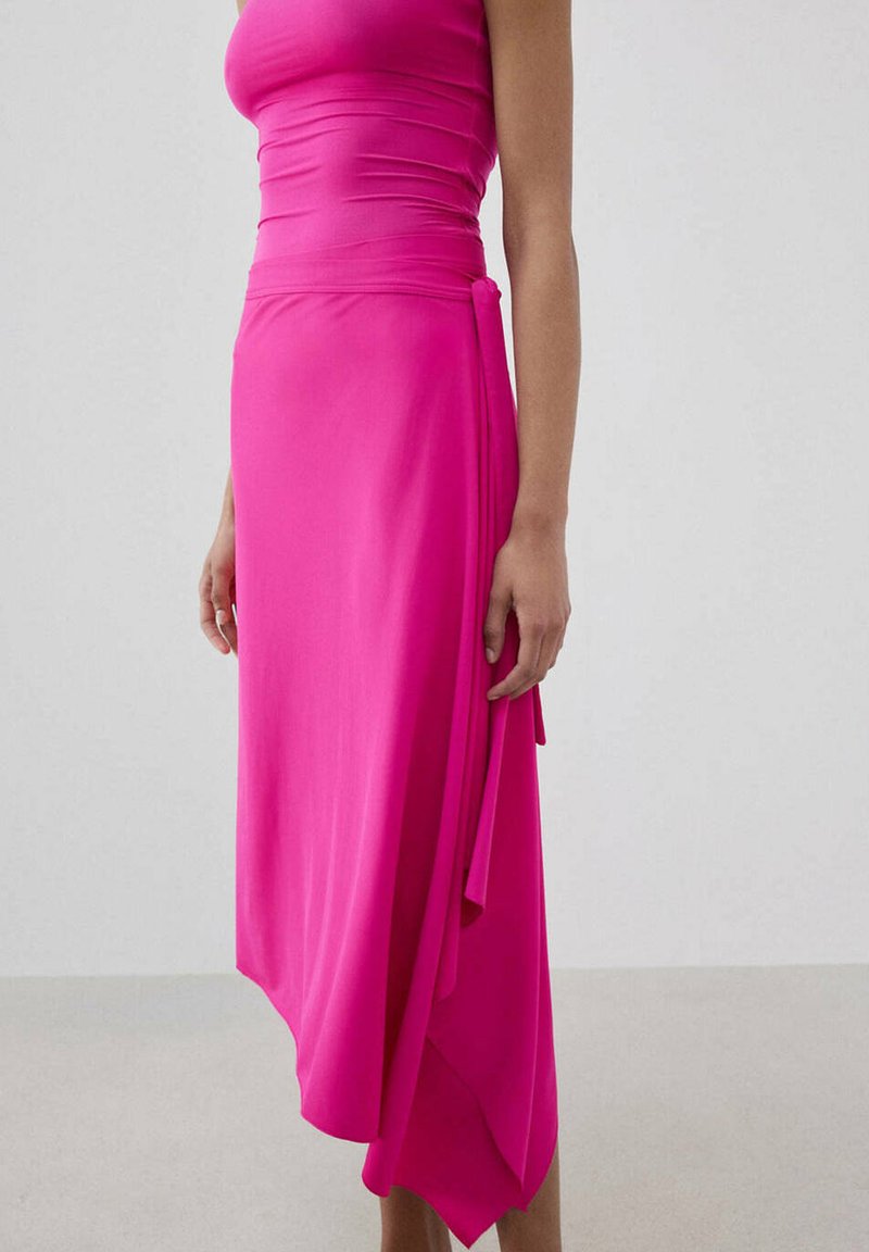 Femme portant un haut sans manches ajusté rose vif et une jupe asymétrique assortie avec un détail de nœud sur le côté, debout devant un fond uni.