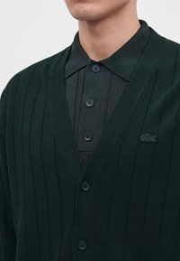 Homme portant un cardigan côtelé vert foncé par-dessus un polo vert foncé boutonné avec col et logo crocodile sur le cardigan.