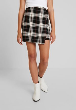 Mujer con falda cruzada de cuadros en negro, blanco y rojo con botones en el lateral, top blanco sin mangas y botines blancos hasta el tobillo.