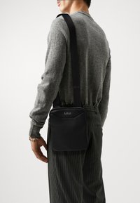 Borsa a spalla in pelle nera con forma rettangolare, chiusura con zip e accentuazione del logo. Il materiale appare liscio con dettagli di cucitura a contrasto.