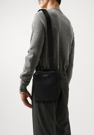 Sac à bandoulière en cuir noir de forme rectangulaire, fermeture zip, et accent logo. Le matériau semble lisse avec des détails de couture contrastés.