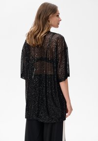 Kobieta z włosami do ramion, nosząca duże kolczyki w kształcie kółek oraz przejrzysty czarny top z cekinami na czarnych spodniach, zwrócona w prawo na białym tle.