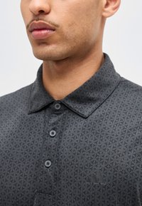 Νεαρός άνδρας που φοράει μαύρο πουκάμισο με σχέδια, γιακά, κουμπιά και λογότυπο Adidas στο αριστερό στήθος.