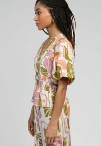 Blouse à motifs floraux avec manches bouffantes, taille cintrée et silhouette fluide dans des couleurs pastel douces, principalement rose, vert et crème.