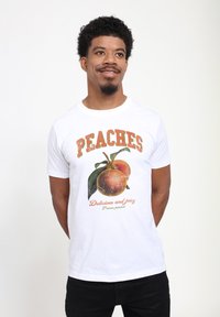 Man som bär en vit t-shirt med en persikografik och texten "PERSIKOR Läckra och saftiga Prunus persica," står med händerna bakom ryggen.