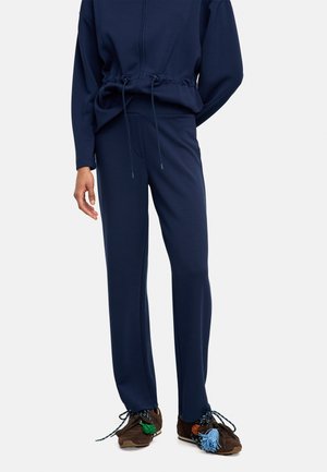 Osoba ubrana w granatowe spodnie na troczki i pasującą bluzę, nosząca ciemnobrązowe trampki ozdobione kolorowymi frędzlami.