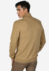 Beiger Rollkragenpullover aus weichem Strickstoff, mit geripptem Kragen und Bündchen sowie geradem Schnitt, getragen zu braunen Hosen.