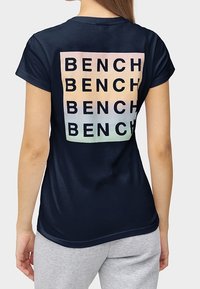 Navy blue t-shirt with short sleeves featuring a large "BENCH" logo in gradient colors on the back. Soft fabric, relaxed fit.