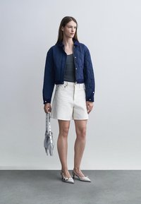 Young woman wearing a navy quilted jacket, gray tank top, white high-waist shorts, white heels, holding a small silver handbag with fringe.