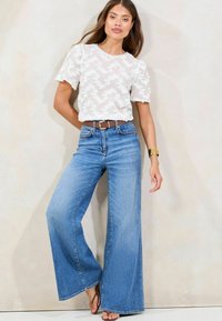 Witte top met korte mouwen en een textuur met bloemmotief; hooggeplaatste lichtblauwe wijde jeans; bruin riem met een gouden gesp; lichte schoenen.