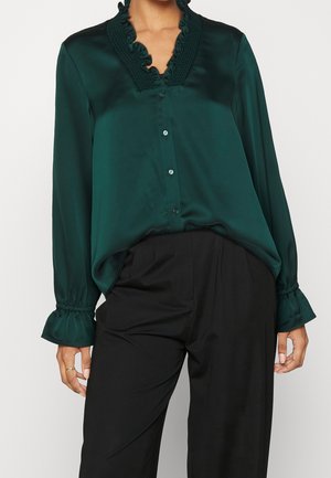 Kobieta w ciemnozielonej satynowej bluzce z falbanianym kołnierzem i mankietami, w połączeniu z czarnymi, dopasowanymi spodniami z wysokim stanem.