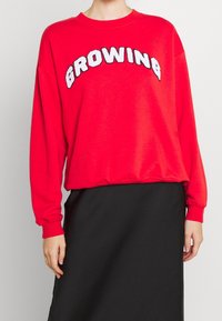 Czerwona bluza z białym napisem "GROWING", długie rękawy i okrągły dekolt. Posiada prążkowane mankiety i luźny krój. Zestawiona z czarną spódnicą.