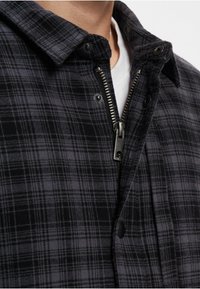 Karo jaka tumši pelēkā un melnā krāsā, ar pogām pie kakla un sudraba rāvējslēdzēju, ar teksturētu apdari un pārbaudītu rakstu.