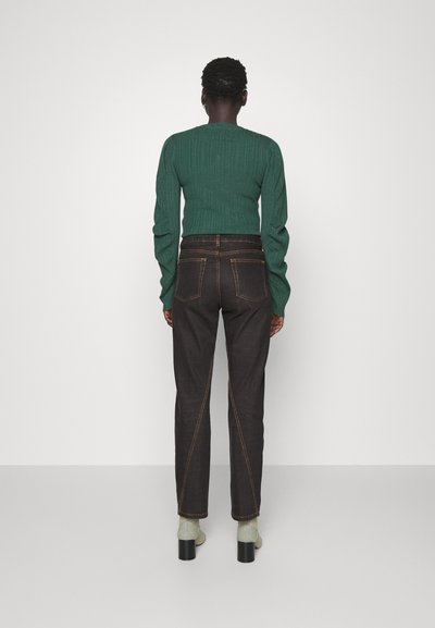 Σκούρο τζιν παντελόνι με διπλή ραφή και μοντέρνα γραμμή, συνδυασμένο με ένα υφασμένο πράσινο πλεκτό πουλόβερ και πράσινες κάλτσες μέχρι τον αστράγαλο.