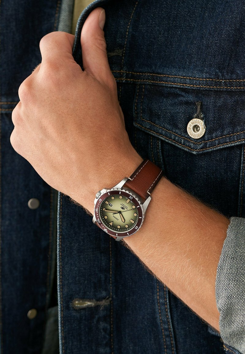 Brauner Lederarmbanduhr mit einem Gehäuse aus silbernem Edelstahl, grünem Zifferblatt und roter Lünette; verfügt über lumineszierende Stundenmarkierungen und ein Datumsfenster.