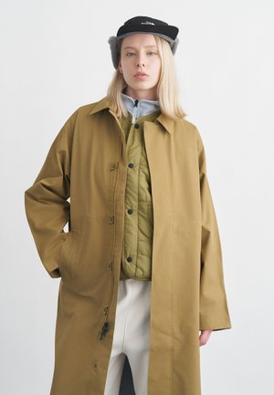 Jeune personne portant un long manteau beige par-dessus une veste matelassée verte, un pantalon clair, un chapeau en polaire gris et une casquette noire, debout devant un fond blanc.