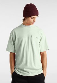 Mintgrünes Baumwoll-T-Shirt mit Rundhalsausschnitt und kurzen Ärmeln, mit einer kleinen Tasche auf der linken Brust. Getragen mit einer burgunderroten Strickmütze.