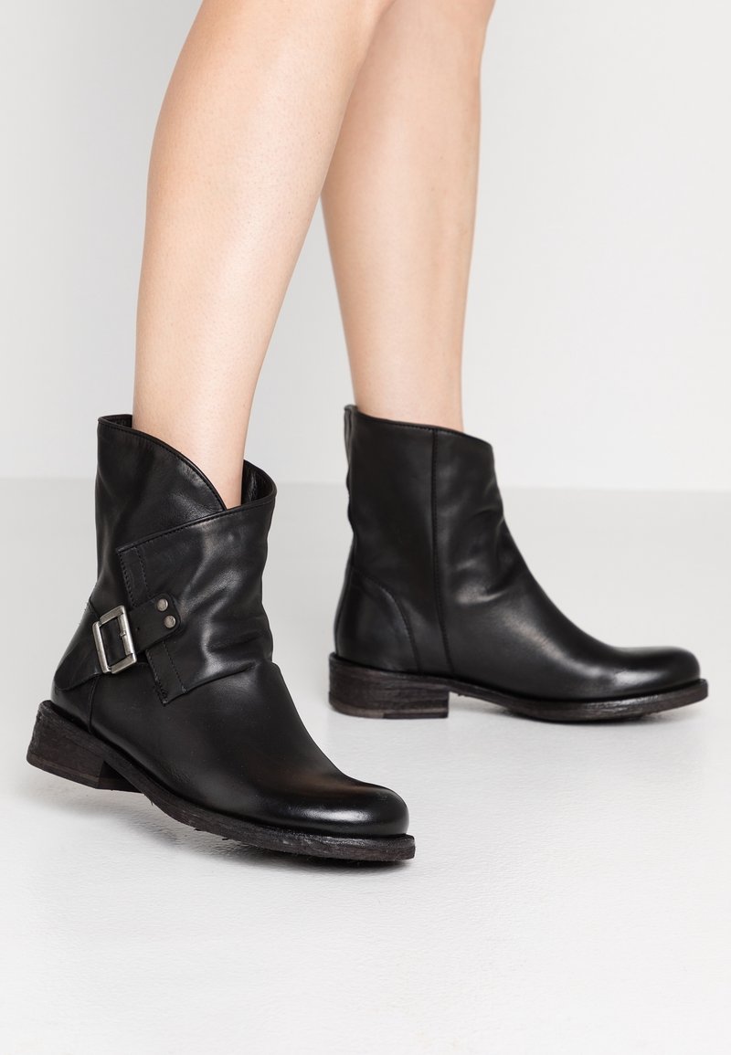 Bottes à chevilles en cuir noir avec une boucle latérale portées sur des jambes nues, dotées de talons bas empilés et d'un design légèrement décontracté.