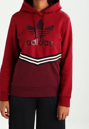 Osoba w dwukolorowej czerwonej bluzie z kapturem Adidas z logo i biało-czarnymi paskami, w czarnych spodniach, stojąca na jednolitym tle.