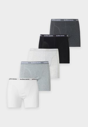 Un homme mince et musclé se tient pieds nus, portant un boxer gris avec une ceinture de marque, sur un fond blanc uni.