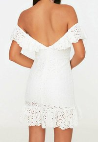 Valkoinen olkaimeton Mekko, jossa on röyhelöhihoja ja tiukka vartalo. Sisältää pitsiyksityiskohtia ja laajenevan, aaltoilevan helman.