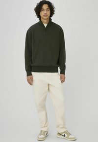 Pull en maille vert foncé avec col à demi-fermeture éclair, associé à un pantalon en velours côtelé beige clair et des baskets beiges avec un design blanc et noir.