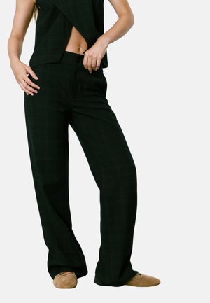 Mujer vestida con pantalones anchos de cuadros negros y una parte superior sin mangas a juego, ajustándose la cinturilla, con zapatillas de color tostado en los pies.