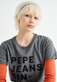 Camiseta gris con texto negro "PEPE JEANS", combinada con una camisa de manga larga naranja por debajo. El modelo lleva una cinta de cuentas en la cabeza.