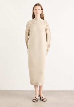 Beige gebreide jurk met een hoge kraag, geribde mouwen en een rechte silhoutte. Afgewerkt met een geribde zoom. Zwarte sandaaltjes met hak en bandjes.