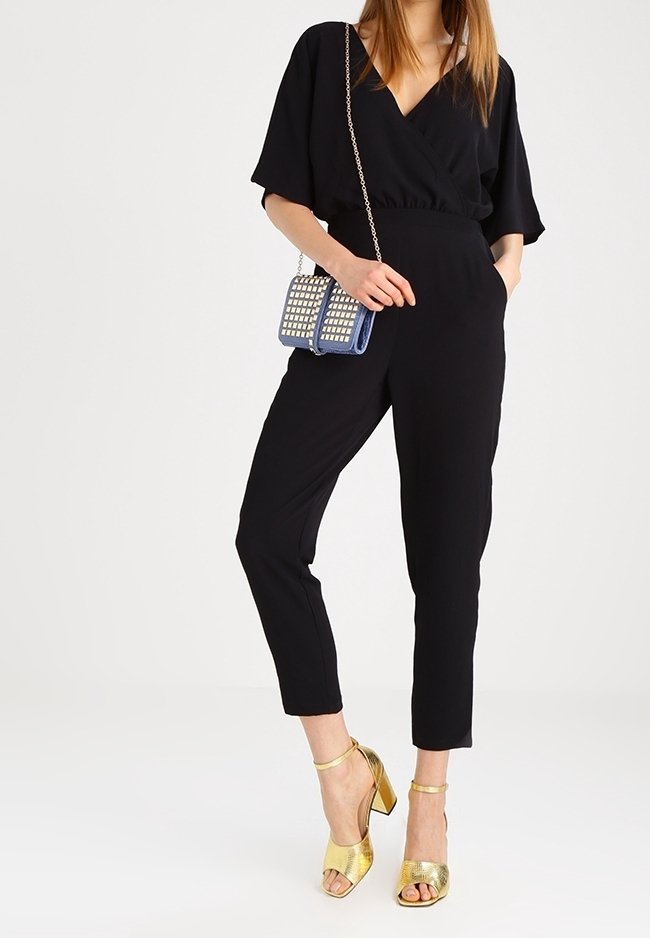 Černý overal s krátkými rukávy a výstřihem do V. Doplněn modrou kabelkou s cvočky a zlatými sandály na podpatku. Hladký povrch látky.