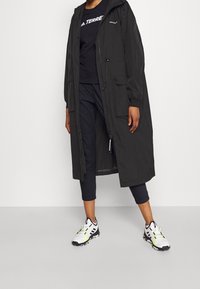 Chaqueta larga negra con capucha y cremallera, llevada sobre una camiseta negra y pantalones azul marino, combinada con zapatillas blancas y negras que tienen acentos en neón.