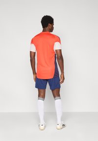 Άνδρας που φοράει κόκκινη και λευκή φανέλα ποδοσφαίρου, μπλε σορτς, λευκές ψηλές κάλτσες και λευκά παπούτσια με τάπες, στέκεται με την πλάτη στραμμένη σε μια απλή φόντο.