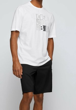 Homme portant un t-shirt blanc à manches courtes avec un motif graphique et un short noir arrivant aux genoux, debout avec une main dans la poche sur un fond uni.