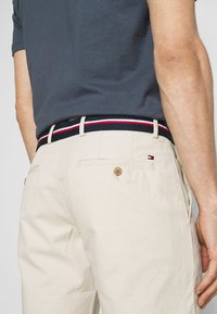 Housut khakia beige-värissä, jossa on tummansininen vyö, jossa on punaisia ja valkoisia päällekkäin kulkevia viivoja. Housuissa on takatasku napilla ja säädettävä vyötärö.