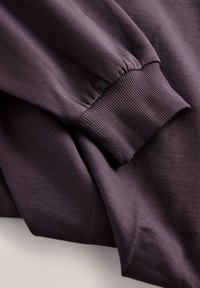Poignet de manche longue avec finition en tricot côtelé sur un vêtement en tissu doux violet foncé, montrant la texture et les plis du tissu.